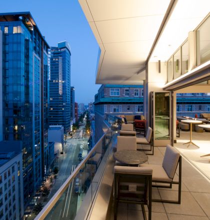 A modern cityscape viewed from a stylish rooftop terrace with seating, tables, and an open indoor area, during evening time with buildings lit up.