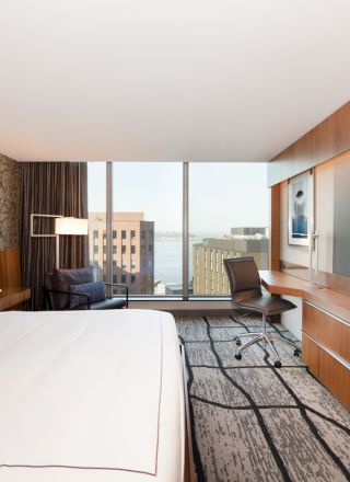 A modern hotel room with a large bed, wall-mounted TV, desk, and chair, featuring floor-to-ceiling windows with a city view in the background.