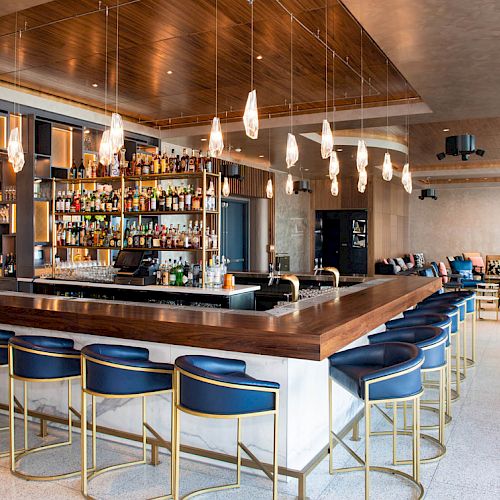 The image shows a modern bar with a wooden counter, blue bar stools, hanging lights, a fully stocked bar, and seating areas in the background.