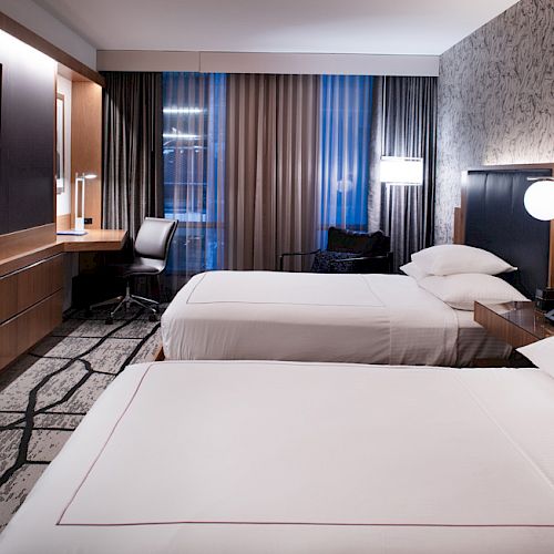 The image shows a modern hotel room with two beds, a wall-mounted TV, a desk with a chair, bedside lamps, and large windows with curtains.