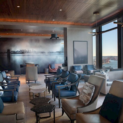 A modern lounge area featuring plush seating, a cityscape mural, large windows with water views, and ambient lighting, creating a relaxed atmosphere.