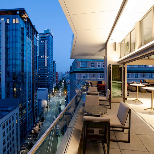 A modern balcony overlooking a cityscape at dusk, featuring tables and chairs near a glass railing with buildings lining the street below.