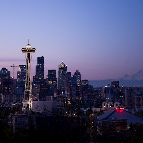 City skyline at dusk featuring a prominent tower with a rotating observation deck, surrounded by diverse architecture and a distant mountain.