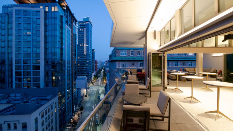 The image shows a modern cityscape with high-rise buildings, viewed from a balcony with tables and chairs, at twilight.