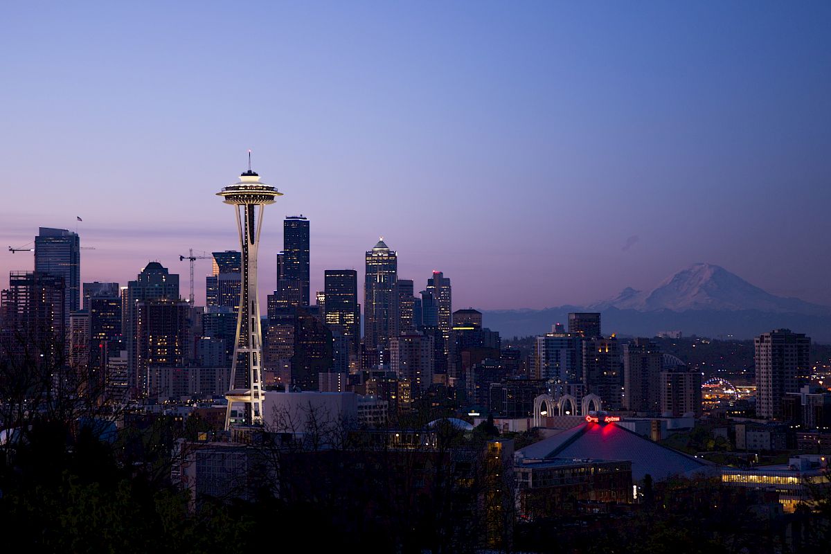 The image shows a cityscape at dusk with prominent skyscrapers, the Space Needle, and a distant mountain.