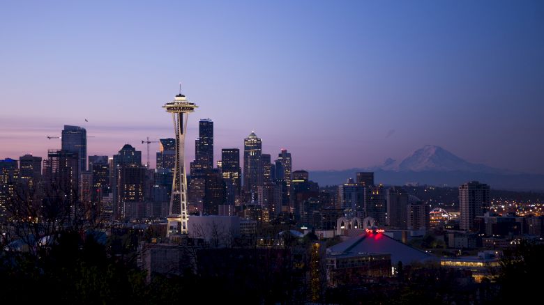 The image shows a cityscape at dusk with prominent skyscrapers, the Space Needle, and a distant mountain.