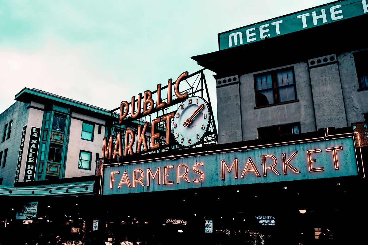 This image shows the neon signs of "Public Market" and "Farmers Market" on building facades, with a clock and part of a hotel visible in the background.