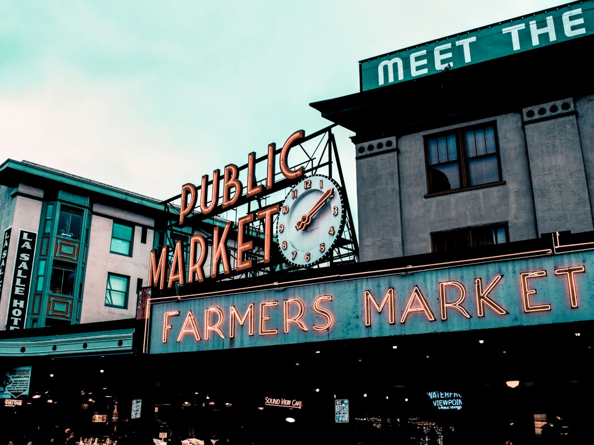 This image shows the neon signs of "Public Market" and "Farmers Market" on building facades, with a clock and part of a hotel visible in the background.