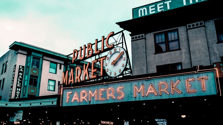 This image shows the neon signs of "Public Market" and "Farmers Market" on building facades, with a clock and part of a hotel visible in the background.