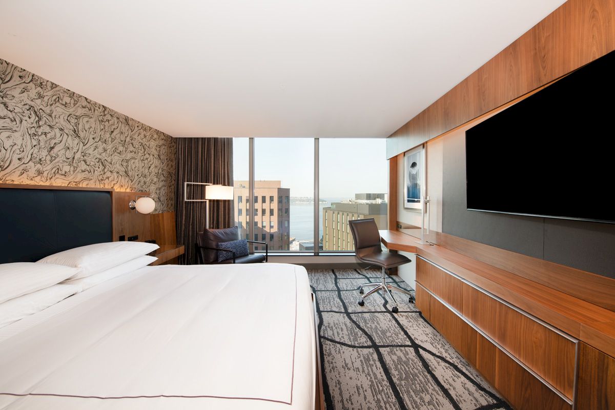 A modern hotel room features a large bed, desk, chair, and TV, with a view of buildings and the sea through a wide window.