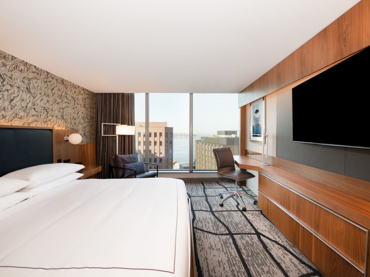 A modern hotel room features a large bed, desk, chair, and TV, with a view of buildings and the sea through a wide window.