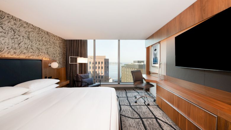 A modern hotel room features a large bed, desk, chair, and TV, with a view of buildings and the sea through a wide window.