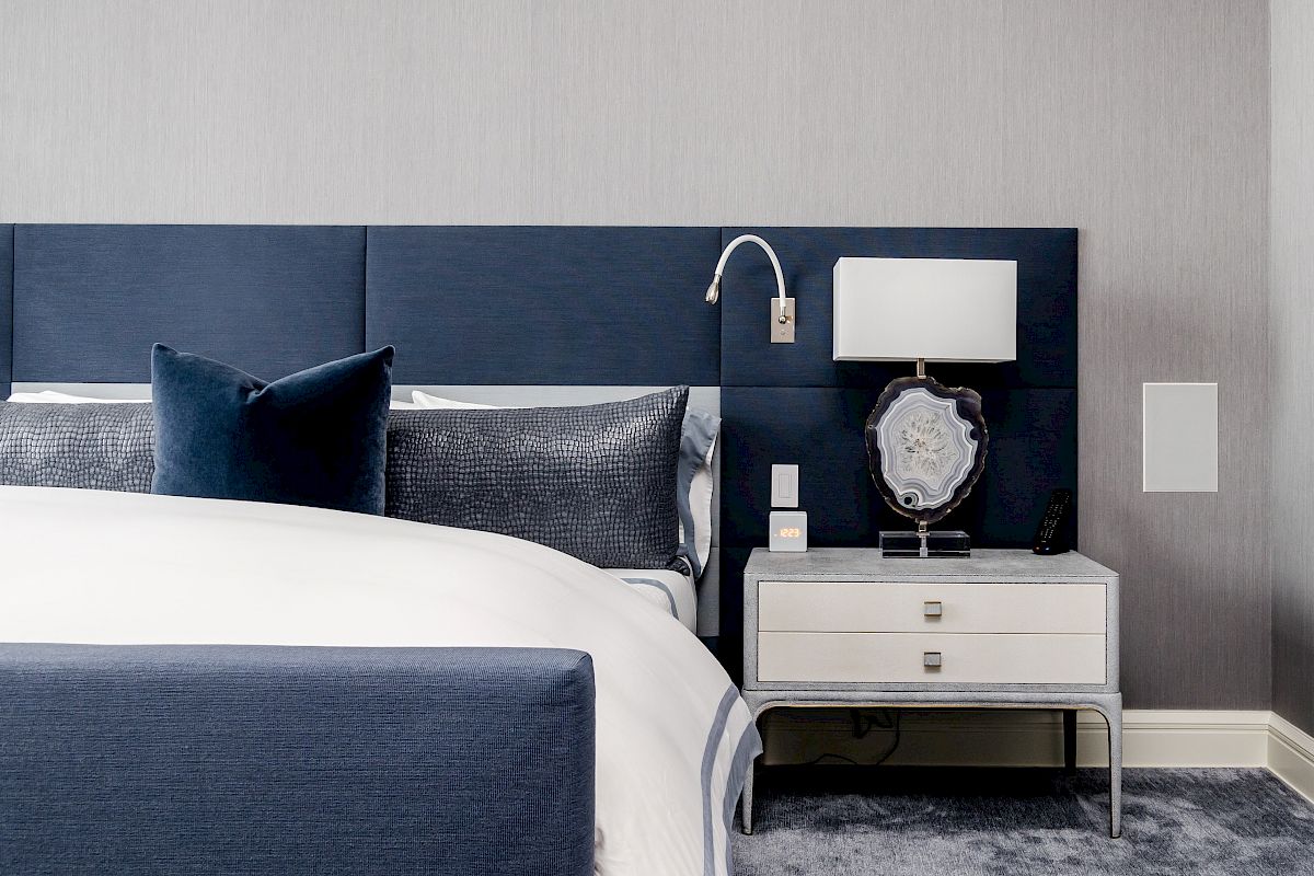 A modern bedroom with a blue and white bed, matching pillows, a bedside table with a lamp, decorative item, and a wall-mounted reading light.
