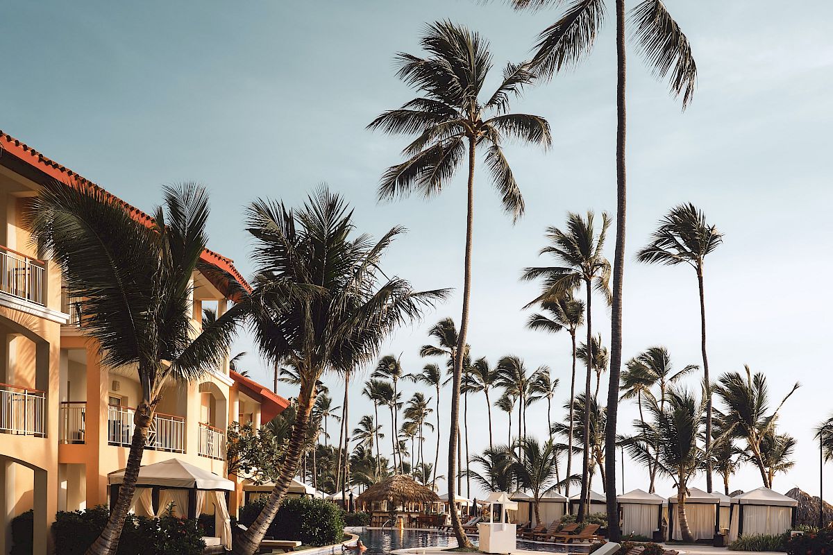 A luxurious resort pool area is surrounded by palm trees and sun loungers, with a multi-story hotel building in the background under a clear sky.