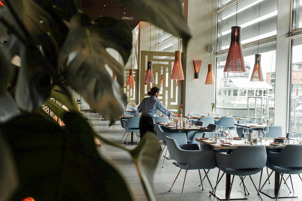 A modern restaurant interior with stylish chairs, pendant lights, large windows, and a server preparing a table next to a leafy plant.