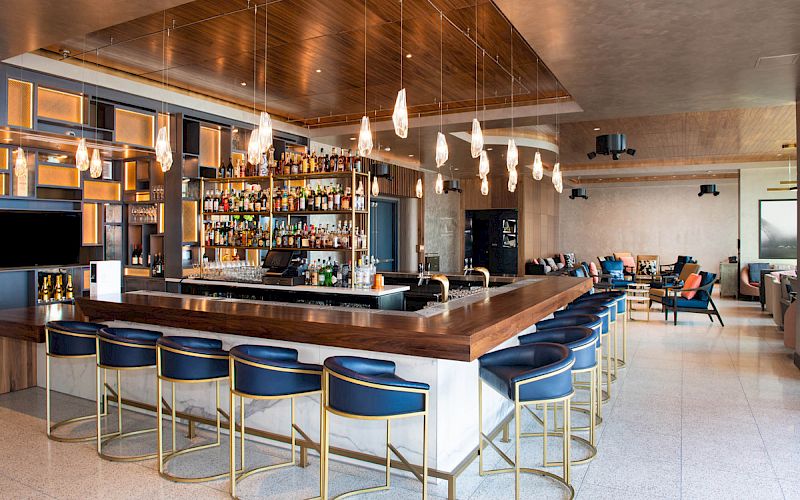 An upscale bar with a wooden counter, blue bar stools with gold frames, hanging pendant lights, and a well-stocked liquor shelf; modern seating area.