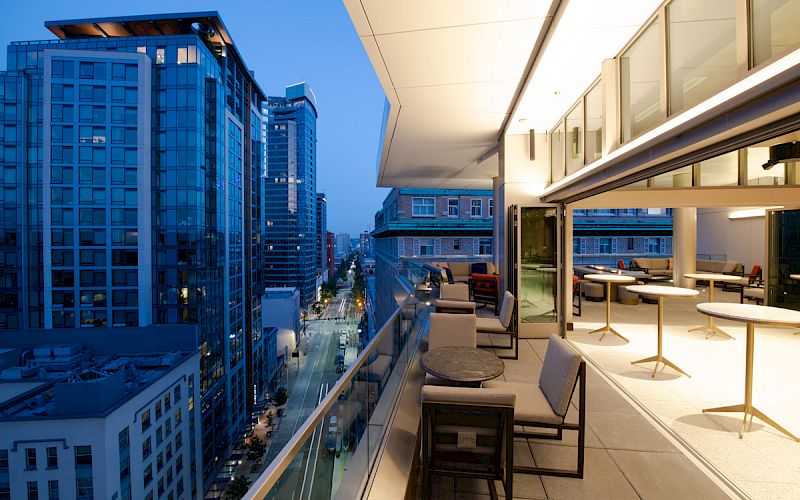 A modern cityscape in the evening featuring tall buildings and a stylish balcony with seating and tables overlooking a busy street below.