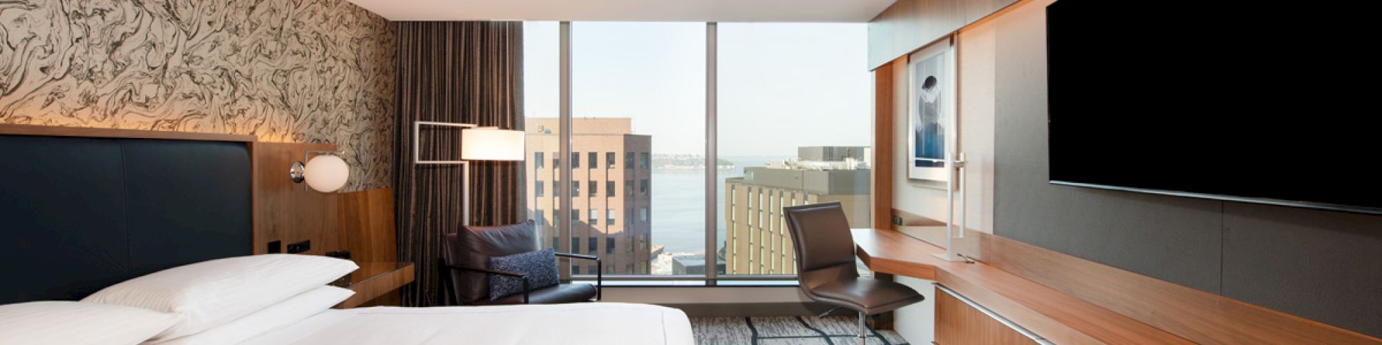 A modern hotel room featuring a large bed, TV, desk, chair, and a window view of city buildings and water in the distance.